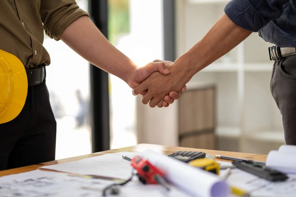 Two construction professionals shaking hands over blueprints and tools